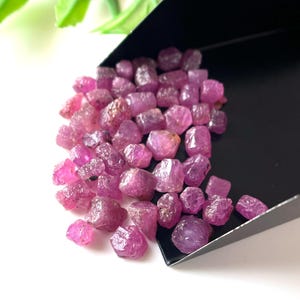 May include: A selection of raw ruby gemstones, in a deep pinkish-red hue. The uncut stones have a slightly translucent quality and are presented on a black tray. The background is white.