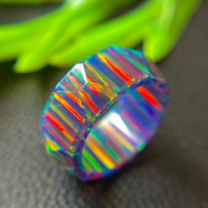 May include: A faceted ring featuring a vibrant, iridescent play of color with vertical bands of bright orange, neon green, and deep blue. The surface of the band is polished and reflects light, showcasing a multi-colored, prismatic pattern throughout.
