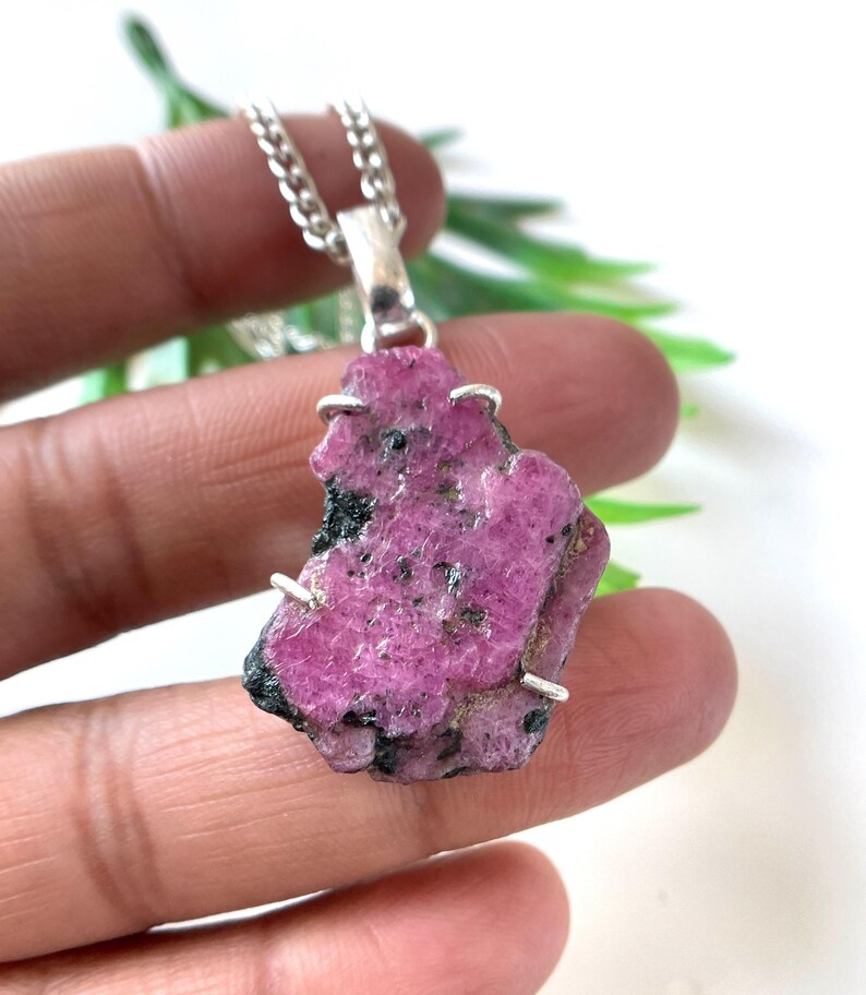 May include: A raw, pink ruby pendant necklace. The irregularly shaped gemstone is a vibrant magenta color with black inclusions. The pendant is attached to a silver chain. The necklace is held in a hand.
