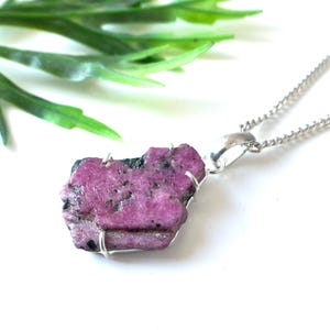 May include: A raw, pink ruby pendant necklace with a silver-colored chain. The ruby is irregularly shaped and has a rough texture. The necklace is displayed on a white surface with green foliage in the background.
