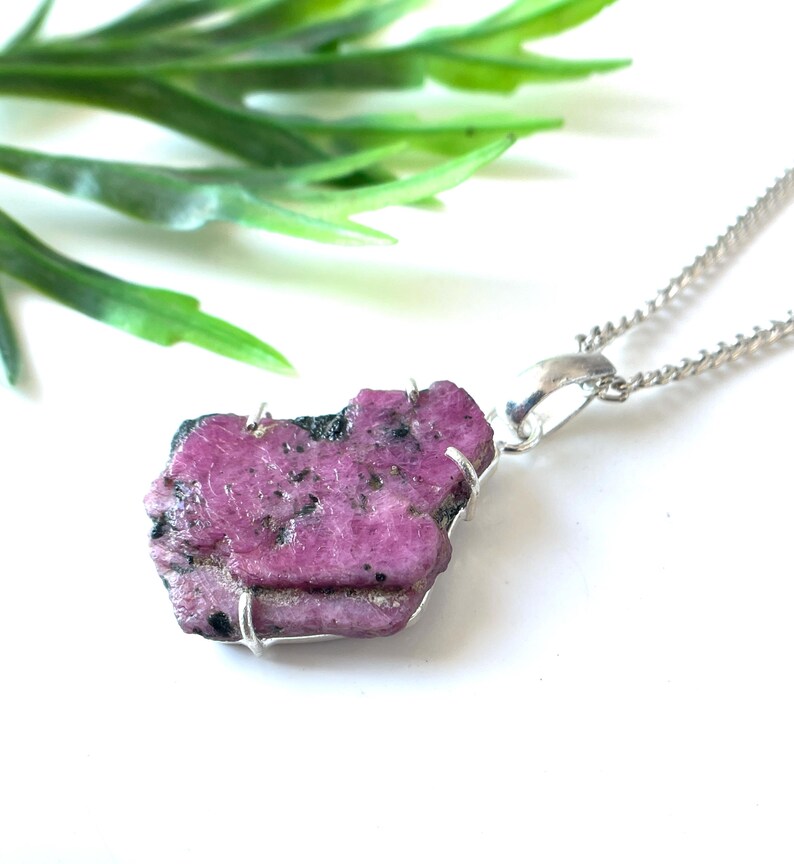 May include: A raw, pink ruby pendant necklace with a silver-colored chain. The ruby is irregularly shaped and has a rough texture. The necklace is displayed on a white surface with green foliage in the background.