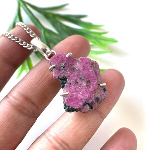 May include: A raw, pink ruby pendant with a silver chain. The ruby has dark black inclusions and is held in place by silver prongs. The pendant is displayed against a white background with green foliage.