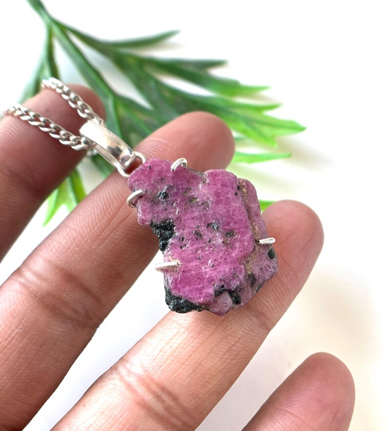 May include: A raw, pink ruby pendant with a silver chain. The ruby has dark black inclusions and is held in place by silver prongs. The pendant is displayed against a white background with green foliage.