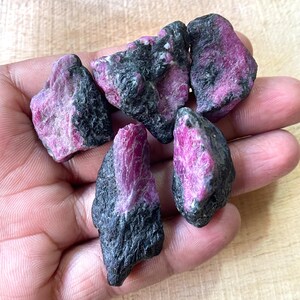 Könnte beinhalten: Eine Sammlung roher Rubin-Edelsteine mit tiefrosa Farbton, kontrastiert durch dunkelgraue Matrix. Die rohen Steine werden in einer Hand gehalten und zeigen ihre natürlichen, unregelmäßigen Formen und Texturen. Diese Edelsteine eignen sich ideal für Schmuckherstellung oder Sammlungen.