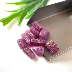 May include: A collection of rough, uncut ruby gemstones in varying sizes and shapes. The gemstones are a deep pinkish-red color and are displayed on a silver tray. A sprig of green leaves is in the background.
