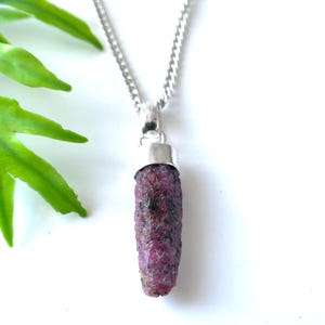 Peut inclure: Un collier en argent avec un pendentif en rubis brut. Le rubis est d'un rouge-rose profond avec une surface textur&eacute;e. Le pendentif mesure environ 2,5 cm de long. Le collier est pr&eacute;sent&eacute; sur fond blanc avec des feuilles vertes.