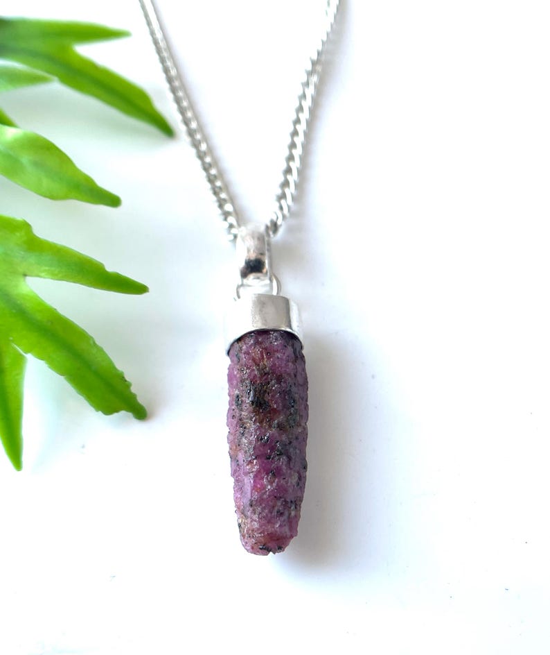 Peut inclure: Un collier en argent avec un pendentif en rubis brut. Le rubis est d'un rouge-rose profond avec une surface textur&eacute;e. Le pendentif mesure environ 2,5 cm de long. Le collier est pr&eacute;sent&eacute; sur fond blanc avec des feuilles vertes.