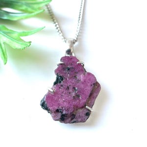 May include: A raw, pink ruby pendant necklace. The irregularly shaped gemstone is mounted on a silver-colored setting and hangs from a delicate chain. The ruby has dark flecks and a rough texture, showcasing its natural beauty.