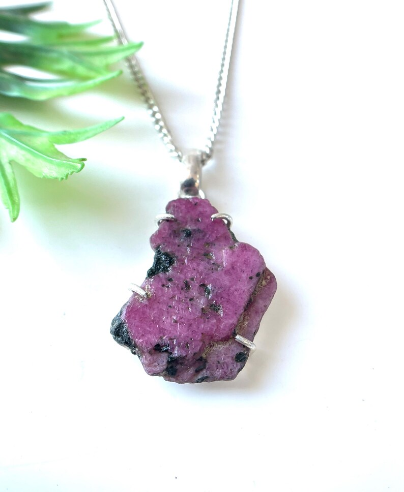 May include: A raw, pink ruby pendant necklace. The irregularly shaped gemstone is mounted on a silver-colored setting and hangs from a delicate chain. The ruby has dark flecks and a rough texture, showcasing its natural beauty.