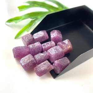 May include: A collection of small, rough-cut, magenta-colored gemstones with a crystalline texture, resting inside a black metal scoop. The stones have irregular, blocky shapes and a semi-translucent appearance with visible surface facets.