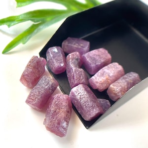 Puede incluir: Una colección de cristales de rubí en bruto, sin pulir, de color púrpura rosado, con una estructura hexagonal natural y superficies texturizadas, que descansan dentro de una pala de metal negro. Las piedras muestran tonos magenta.