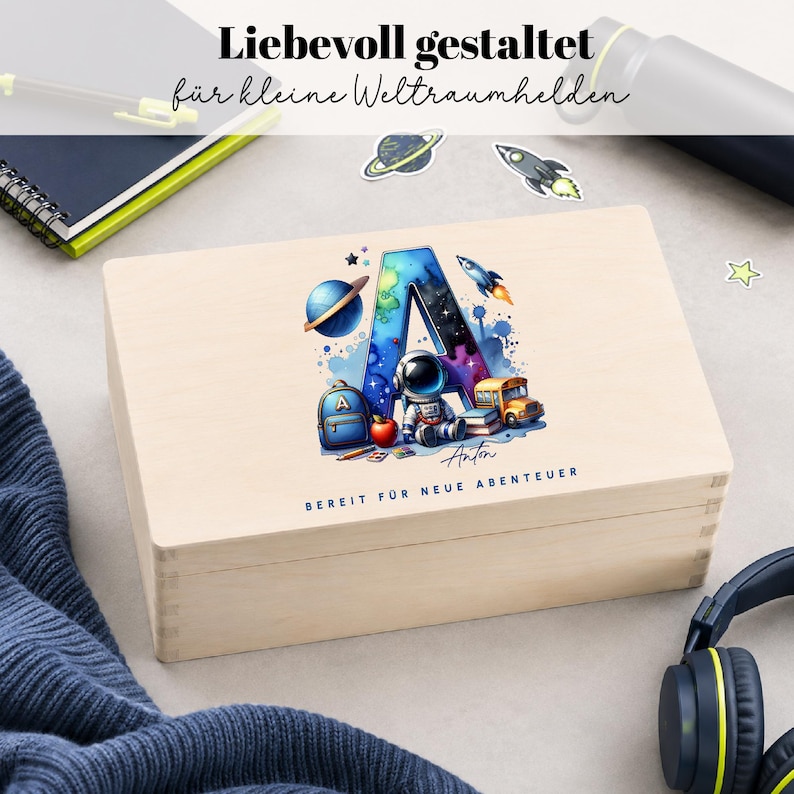 Peut inclure: Un coffret en bois clair avec une illustration color&eacute;e sur le th&egrave;me de l'espace. Le design comprend la lettre "A" remplie d'une sc&egrave;ne de galaxie, un astronaute, un bus scolaire et le nom "Anton". Le coffret porte l'inscription "BEREIT F&Uuml;R NEUE ABENTEUER" en bas.