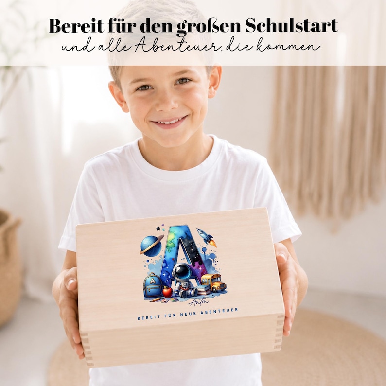 Peut inclure: Une bo&icirc;te en bois avec un design color&eacute; sur le th&egrave;me de l'espace, avec la lettre "A", une plan&egrave;te, une fus&eacute;e et un astronaute. La bo&icirc;te porte les inscriptions "Bereit f&uuml;r den gro&szlig;en Schulstart und alle Abenteuer. die kommen" et "Bereit f&uuml;r neue Abenteuer".