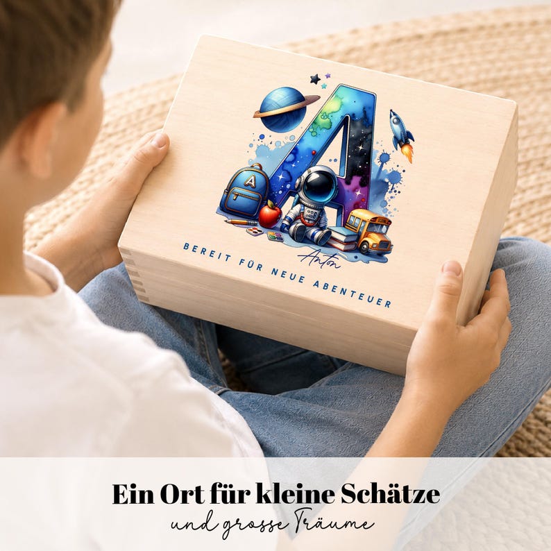 Peut inclure: Un coffret en bois avec un design color&eacute; sur le th&egrave;me de l'espace. Le motif comprend une grande lettre "A" remplie d'un motif de galaxie, une plan&egrave;te, une fus&eacute;e et un astronaute. Le coffret porte l'inscription "BEREIT F&Uuml;R NEUE ABENTEUER" et "Anton".