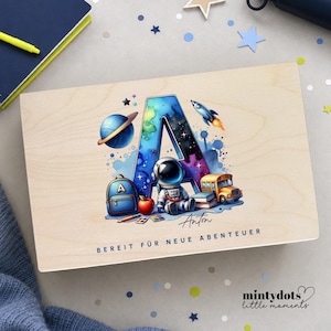 Peut inclure: Bo&icirc;te en bois avec une illustration color&eacute;e repr&eacute;sentant la lettre "A" dans un style aquarelle, sur le th&egrave;me de l'espace, et le nom "Anton". Le design comprend une plan&egrave;te, une fus&eacute;e, un astronaute, un bus scolaire et un sac &agrave; dos. Le texte "BEREIT F&Uuml;R NEUE ABENTEUER" est en bas.