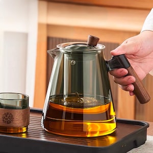 May include: A glass teapot with a wooden handle and lid, filled with amber liquid. The teapot sits on a black tray, next to a glass with a wooden sleeve. The teapot has a built-in infuser. The handle is dark brown.