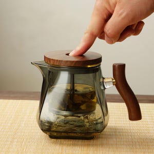 Könnte beinhalten: Eine Glasteekanne mit Holzdeckel und Griff. Die Teekanne hat eine dunkelgraue Tönung und enthält Teeblätter. Der Griff ist gebogen und aus Holz. Der Deckel ist rund und ebenfalls aus Holz.