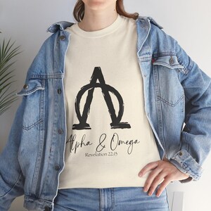 May include: Cream-colored t-shirt featuring a black graphic of the Greek letters Alpha and Omega. Below the graphic, the words "Alpha & Omega" are written in a script font, with "Revelation 22:13" underneath.