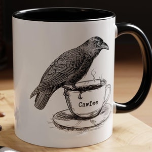Op de afbeelding: Witte keramische mok met een zwarte rand en handvat. De mok heeft een zwart-witte illustratie van een raaf die op een kopje koffie zit met het woord "Cawfee" erop.