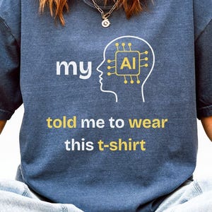 Könnte beinhalten: Ein dunkelblaues T-Shirt mit dem Text "my AI told me to wear this t-shirt" in Weiß und Gold. Das Design zeigt ein stilisiertes Gehirn mit einer KI-Leiterplatte. Teil einer Bekleidungskollektion.
