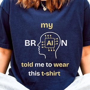 Könnte beinhalten: Marineblaues T-Shirt mit goldenem Text "my BRAIN told me to wear this t-shirt". Das Design zeigt eine weiße Umrisszeichnung eines Kopfes mit einer KI-Leiterplatte. Teil einer Bekleidungskollektion.