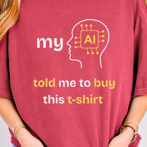May include: Crimson-colored t-shirt with the text "my AI told me to buy this t-shirt." The design features a white outline of a head with an AI chip graphic. The model is wearing blue jeans.
