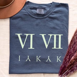 May include: A navy blue t-shirt with the Roman numerals VI VII and the letters IΛKAKK printed in light green. A brown hat and a bundle of dried flowers are in the background. The word "DENIM" is printed in the lower left corner.