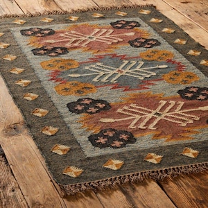 Könnte beinhalten: Rechteckiger Teppich mit geometrischem Muster in Blau-, Braun-, Rot- und Gelbtönen. Der Teppich hat einen dunkelgrauen Rand mit rautenförmigen Akzenten und Fransen. Das Design zeigt stilisierte Blumen- und geometrische Formen.