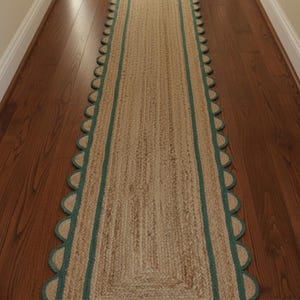 May include: A rectangular jute runner rug with a scalloped edge and a teal border. The rug is placed on a hardwood floor and is designed for a hallway or entryway. The natural fibers and teal accents create a neutral, yet stylish look.