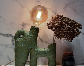 May include: A distinctive green lamp with a textured finish and a large, round lightbulb. The lamp has an abstract, sculptural form. A small vase with dried flowers and a dark glass jar with a cork lid are next to the lamp.