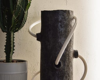 May include: A dark gray cylindrical lamp with a textured surface and white tubing accents. The lamp is on a wooden surface next to a potted cactus. The lamp has a unique, modern design.