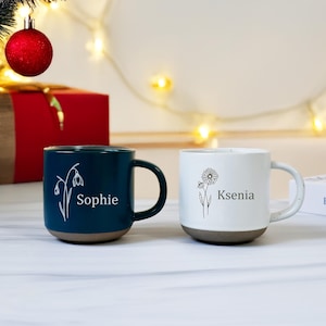 Può includere: Due tazze in ceramica, una verde acqua e una bianca, con disegni floreali e nomi. La tazza verde acqua ha il nome "Sophie", e la tazza bianca ha il nome "Ksenia". Entrambe le tazze hanno una base marrone e manici neri.