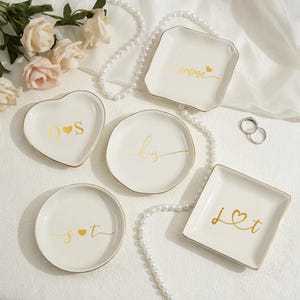 Könnte beinhalten: Eine Sammlung weißer Keramik-Ringschalen in verschiedenen Formen, darunter Herz, rund und quadratisch. Jede Schale hat einen goldenen Rand und goldene Schriftzüge mit Namen und Initialen. Eine Perlenkette und Ringe sind ebenfalls vorhanden.