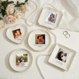 May include: A selection of cream-coloured ceramic trinket dishes in various shapes, including heart, round, and square. Each dish features a photograph and gold trim. Some dishes have text, and a pearl necklace and flowers are in the background.