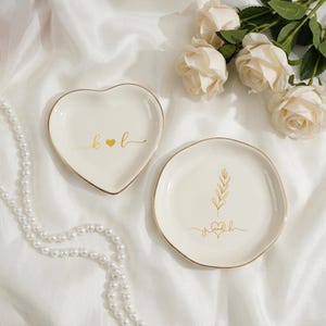 Peut inclure: Deux vide-poches en céramique blanc cassé avec bordure dorée. L'un est en forme de cœur avec "k ♥ l" en script doré. L'autre est rond avec un motif floral doré et "je ♥ h" en script doré. Un collier de perles et des roses en arrière-plan.