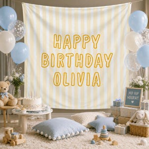 May include: A large fabric backdrop featuring vertical pale yellow and white stripes with the text HAPPY BIRTHDAY OLIVIA in bold yellow letters. The scene includes light blue and white balloons, a birthday cake, and plush toys.
