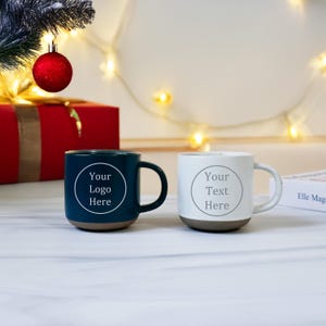 Puede incluir: Dos tazas de cerámica, una verde azulado y otra blanca, con bases y asas marrones. Cada taza tiene un círculo blanco con el texto "Your Logo Here" y "Your Text Here". Las tazas están sobre una superficie blanca con decoraciones navideñas en el fondo.
