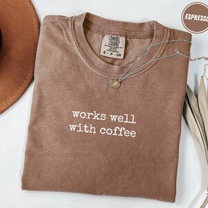 Puede incluir: Camiseta color espresso con el texto blanco "works well with coffee". La camiseta tiene cuello redondo y una pequeña etiqueta cerca del cuello. Un collar plateado con un colgante está drapeado sobre la camiseta.