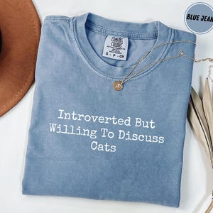 May include: A blue jean coloured t-shirt with the text "Introverted But Willing To Discuss Cats" in white. A gold necklace with a pendant is draped across the shirt. The shirt is made of a soft material.