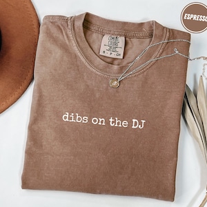 Könnte beinhalten: Espressofarbenes T-Shirt mit dem weißen Schriftzug "dibs on the DJ". Das Shirt hat einen Rundhalsausschnitt und ein kleines Etikett am Kragen. Eine silberne Halskette mit Anhänger liegt über dem Shirt. Ein brauner Hut befindet sich oben links.