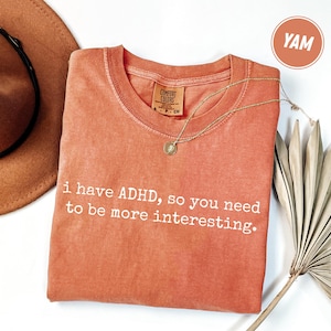 May include: A coral-colored t-shirt with the text "i have ADHD, so you need to be more interesting." A gold necklace with a pendant is draped across the shirt. A brown hat is in the upper left corner.