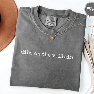 Può includere: Una maglietta grigia Comfort Colors con la scritta bianca "dibs on the villain". Una collana d'argento con un piccolo ciondolo è sulla maglietta. Un cappello marrone e foglie secche sono sullo sfondo.