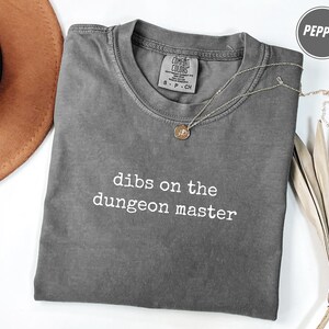 Puede incluir: Una camiseta gris Comfort Colors con el texto "dibs on the dungeon master" en blanco. Un collar plateado con un colgante está sobre la camiseta. Un sombrero marrón y hojas secas están en el fondo.