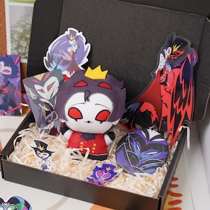 Op de afbeelding: Een zwarte geschenkdoos gevuld met papiersnippers met een pluche pop met een gele kroon, rood-zwarte kleding en rode ogen, omringd door diverse stickers en een ronde button met een gestileerde uilfiguur.