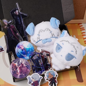 Op de afbeelding: Een verzameling Devil May Cry-merchandise, waaronder twee witte pluche figuren met blauwe oren, twee ronde buttons, twee rechthoekige acryl sleutelhangers en drie uitgestanste stickers die de personages uit de serie afbeelden.