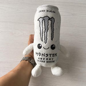 May include: A white plush toy shaped like an energy drink can with small arms and legs. The front features an embroidered black claw logo, a smiling face, and the text ZERO SUGAR MONSTER ENERGY ULTRA WHITE in black thread on the soft fabric.