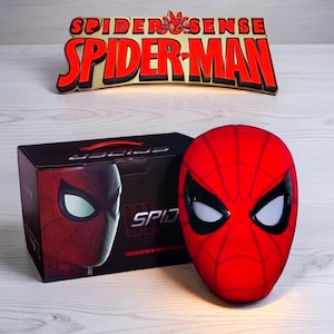 Puede incluir: Una lámpara con forma de máscara de Spider-Man en rojo y negro con lentes de malla blanca, situada junto a una caja negra con un gráfico de Spider-Man. Encima hay un letrero dorado y rojo con el texto SPIDER-SENSE SPIDER-MAN en letras negritas.