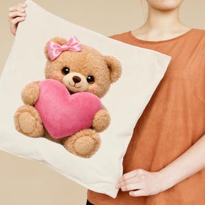 Op de afbeelding: Een beige kussen met een pluche teddybeer die een roze hart vasthoudt. De teddybeer heeft een roze strik en grote, donkere ogen. Het kussen is vierkant en lijkt gemaakt van een zachte stof.