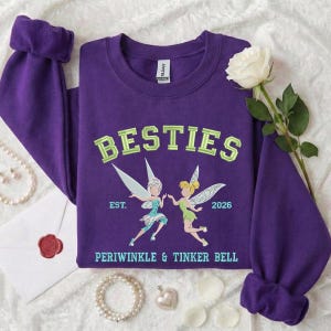 Pode incluir: Um moletom roxo com a palavra "BESTIES" em letras verdes. Abaixo da palavra estão duas fadas de desenho animado, Periwinkle e Tinker Bell, com o texto "EST. 2026" e "PERIWINKLE & TINKER BELL". As mangas são dobradas.