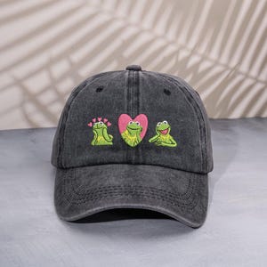 Könnte beinhalten: Dunkelgraue Baseballkappe mit einem gestickten Design mit drei grünen Fröschen. Der mittlere Frosch befindet sich in einem rosa Herzen. Die anderen beiden Frösche befinden sich auf beiden Seiten des Herzens.
